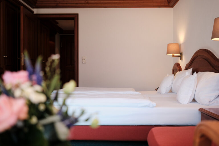 Das gemütliche Hotelzimmer ist mit einem weiß bezogenen Doppelbett, zwei Lampen und Nachttischen ausgestattet, im Vordergrund steht ein Blumenstrauß.