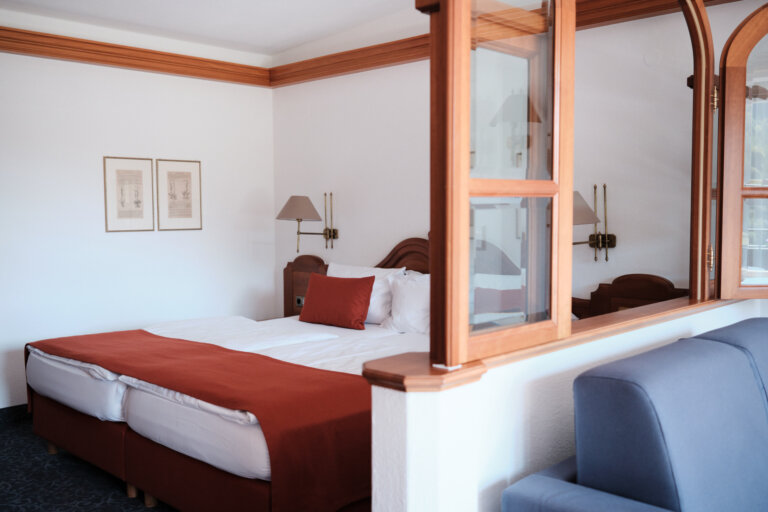 Ein Hotelzimmer mit einem Doppelbett, weißen Bettlaken und einem roten Kissen. An den Wänden hängen zwei Wandleuchten. Es gibt eine Trennwand aus Holz und Glas, die wie ein geöffneter Fensterrahmen aussieht.