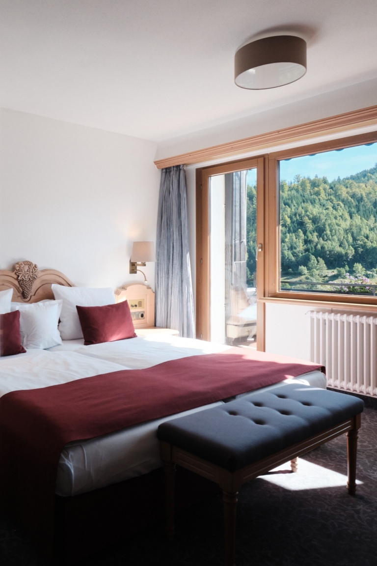 Ein Hotelzimmer mit einem Doppelbett, einer Bank am Fußende und einem großen Fenster mit Blick ins Grüne.