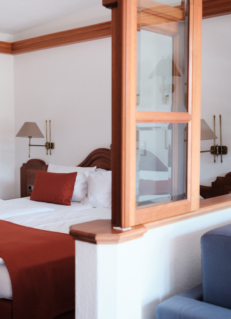 Ein Schlafzimmer mit einem Doppelbett, weißen Bettlaken und einem roten Kissen. An den Wänden hängen zwei Wandleuchten, es gibt eine Trennwand aus Holz und Glas.