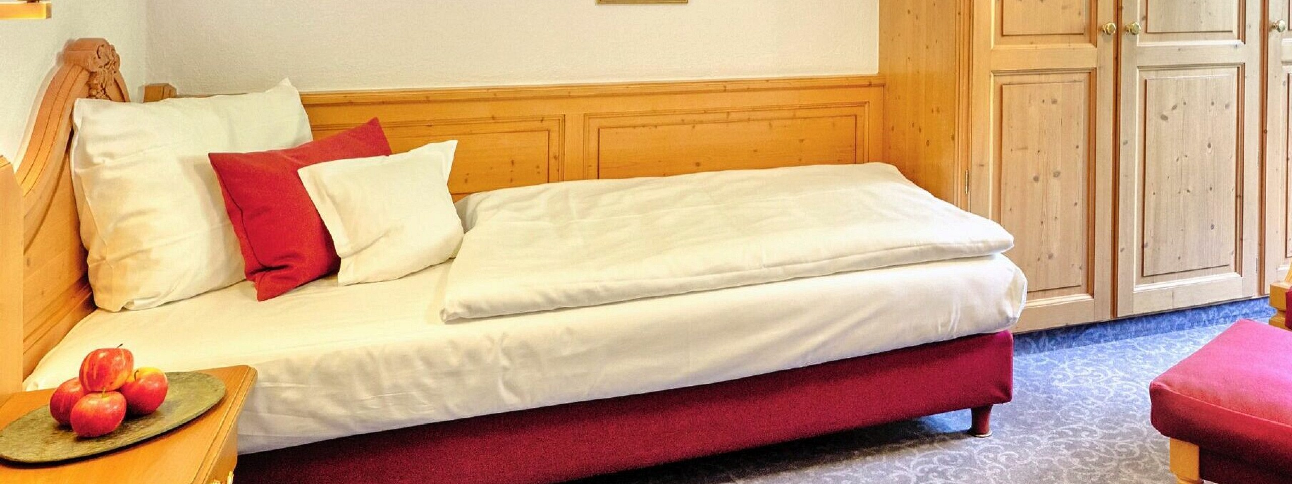 Single-Bett mit rotem Sessel und hölzernem Kleiderschrank im Hotelzimmer im Schwarzwald.